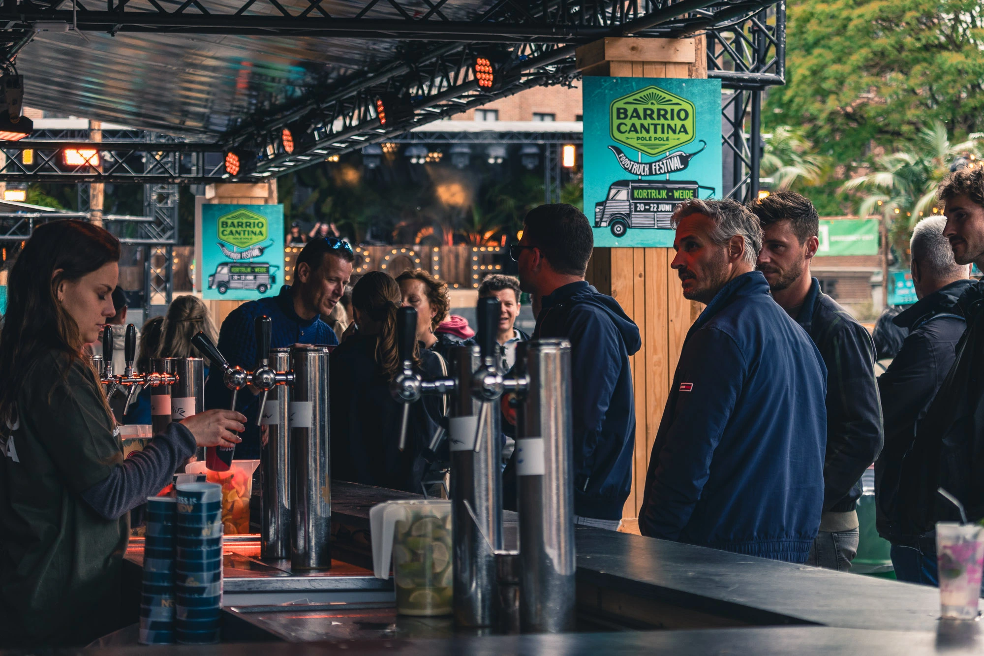 Mensen staan in de rij aan een buitenbar terwijl een barman drankjes serveert; boven de bar zijn borden met Barrio Cantina zichtbaar.