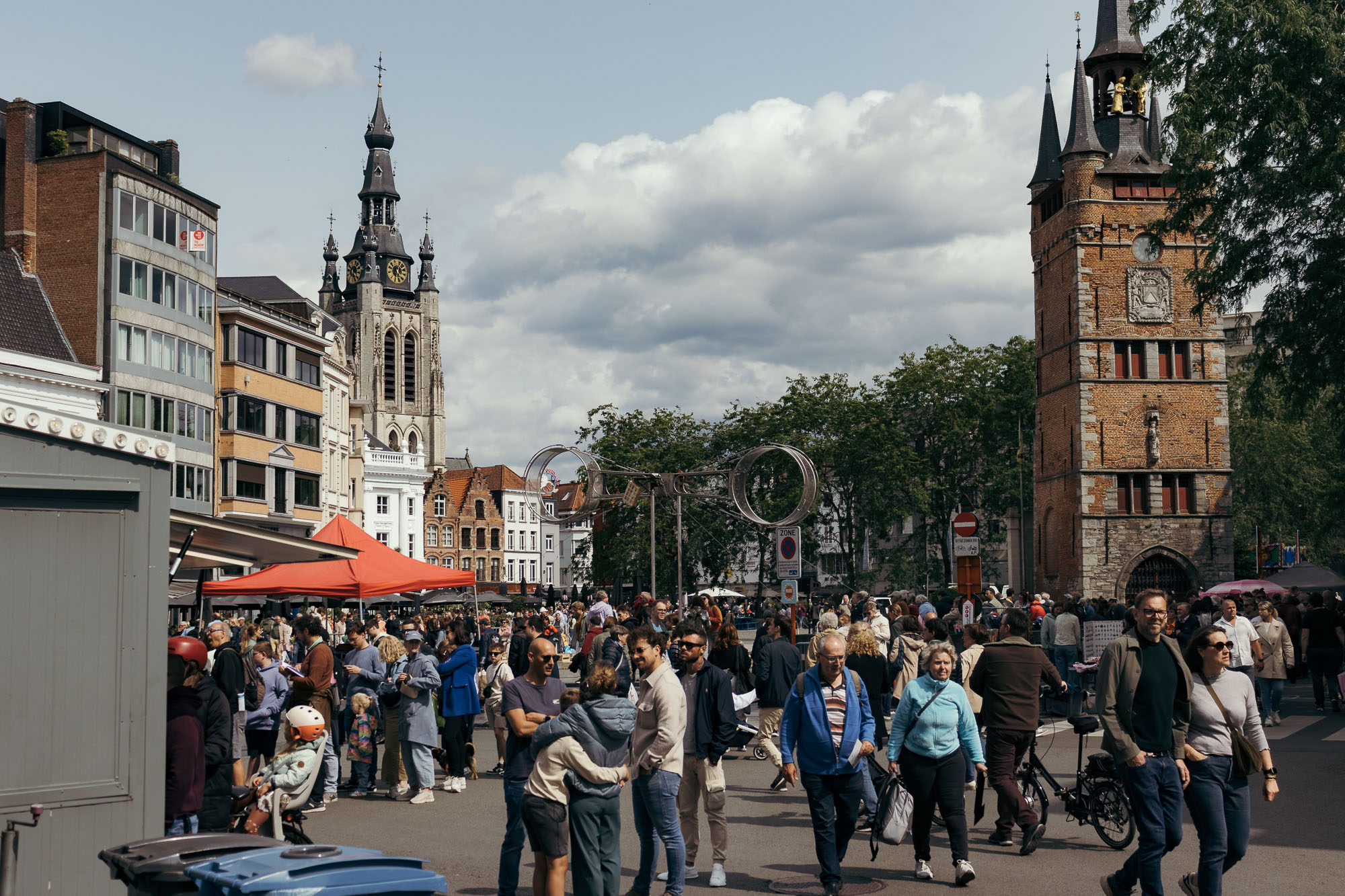 Een bruisende straatscène tijdens Sinksen in Kortrijk, met een diverse menigte in de buurt van historische gebouwen zoals het Belfort en een kerk. Op de achtergrond zijn de metalen hoepels van acrobatische voorstellingen te zien. De sfeer is levendig en sociaal onder een bewolkte hemel.