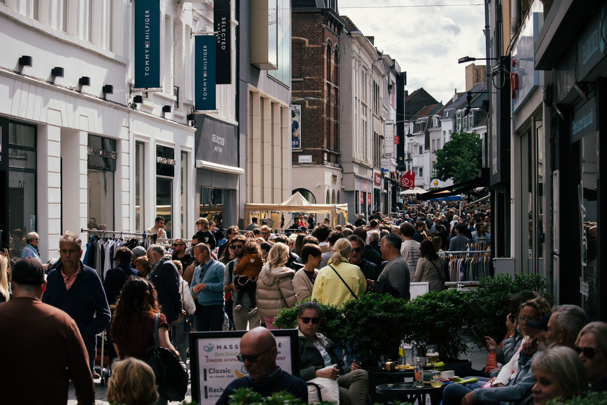 Drukke winkelstraat tijdens Sinksen in Kortrijk, gevuld met een diverse menigte. Mensen snuffelen tussen kledingrekken buiten de winkels onder een heldere hemel. Levendige, bruisende sfeer.