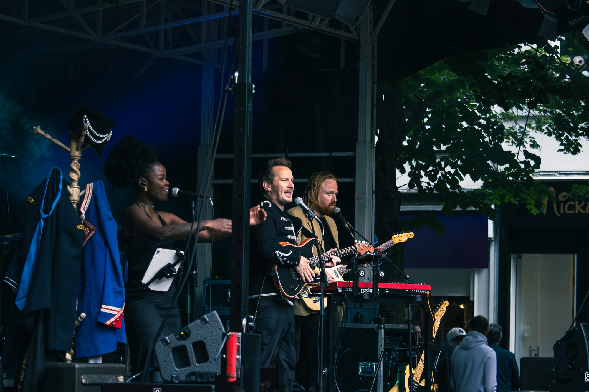 Een energieke band treedt op op een buitenpodium. Een vrouw zingt met enthousiasme, twee mannen spelen op elektrische gitaren. Op een rek hangen kleurrijke jassen. Op de achtergrond staan bomen en toeschouwers.
