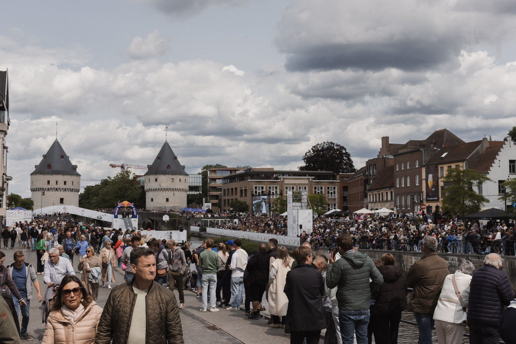 Een grote menigte verzamelt zich tijdens Sinksen in Kortrijk bij een evenement aan de rivier onder bewolkte luchten. De historische Broeltorens en moderne gebouwen zijn op de achtergrond zichtbaar, wat zorgt voor een levendige sfeer.